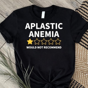 Può includere: T-shirt nera con la scritta bianca "APLASTIC ANEMIA". Sotto, una valutazione con una stella d'oro e quattro stelle vuote, con le parole "WOULD NOT RECOMMEND" sotto.