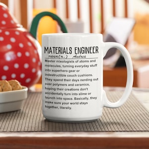 Könnte beinhalten: Eine weiße Keramiktasse mit einem schwarz-weißen Design, auf dem steht "MATERIALS ENGINEER noun(n.) Andrea Master mixologists of atoms and molecules, turning everyday stuff into superhero gear or indestructible couch cushions. They spend their days nerding out over polymers and ceramics, hoping their creations don't accidentally turn into slime or launch into space. Basically, they make sure your world stays together, literally."