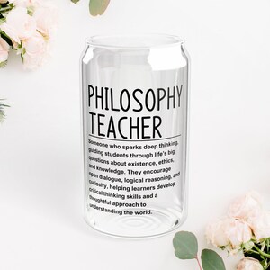 May include: A clear glass can-shaped drinking glass with the words "PHILOSOPHY TEACHER" in black. Below the text is a definition of a philosophy teacher. The glass is surrounded by white flowers and greenery.