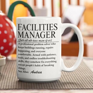Può includere: Una tazza in ceramica bianca con testo stampato in nero che definisce un responsabile delle strutture come un risolutore di problemi professionale che mantiene gli edifici operativi, terminando con la frase See Also: Andrea in corsivo.