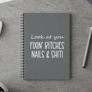May include: A grey spiral-bound notebook featuring the white text "Look at you FIXIN' BITCHES NAILS & SHIT!". The notebook is on a grey surface, with a pen, a computer mouse, and binder clips visible.