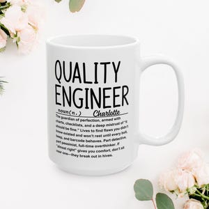 Może przedstawiać: Biały ceramiczny kubek z czarnym napisem "QUALITY ENGINEER", wraz z definicją i imieniem "Charlotte". Kubek ma duży uchwyt i jest otoczony kwiatami.