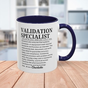 May include: White ceramic mug with a navy blue interior and handle. The mug features the text "VALIDATION SPECIALIST" in black, along with a definition and the name "Charlotte". The mug is on a wooden surface.