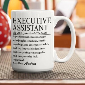 Op de afbeelding: Een witte keramische mok met zwarte bedrukte tekst die een Executive Assistant definieert als een professionele chaosmanager die jongleert met schema's, e-mails, vergaderingen en noodgevallen terwijl onmogelijke deadlines beheersbaar lijken. Bevat de tekst See Also: Andrea.