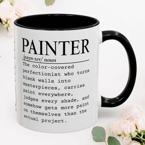 Könnte beinhalten: Ein weißer Keramikbecher mit schwarzem Innenraum und Henkel. Auf der Vorderseite befindet sich ein schwarzer Text, der das Wort PAINTER als einen farbverschmierten Perfektionisten definiert, der leere Wände in Meisterwerke verwandelt.