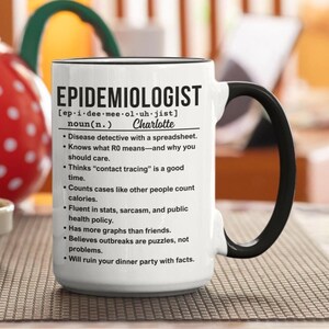 Könnte beinhalten: Weißer Keramikbecher mit schwarzem Henkel und Rand. Der Becher zeigt das Wort "EPIDEMIOLOGIST" in großen schwarzen Buchstaben, zusammen mit einer Definition und dem Namen "Charlotte". Der Becher listet auch Eigenschaften eines Epidemiologen auf.