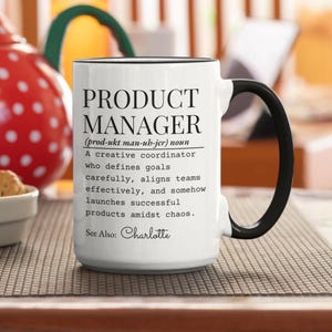 Op de afbeelding: Witte keramische mok met zwarte handgreep en rand. De mok toont de tekst "PRODUCT MANAGER" met een definitie en de naam "Charlotte". Een cadeau voor een productmanager.