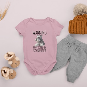 May include: Pink baby onesie with a black graphic that says "WARNING PROTECTED BY SCHNAUZER" and a picture of a miniature schnauzer. The onesie is laid out on a white surface with a pair of brown baby booties, a pair of gray sweatpants, and a brown knit hat with a white pom-pom.