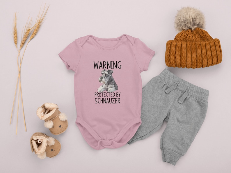 May include: Pink baby onesie with a black graphic that says "WARNING PROTECTED BY SCHNAUZER" and a picture of a miniature schnauzer. The onesie is laid out on a white surface with a pair of brown baby booties, a pair of gray sweatpants, and a brown knit hat with a white pom-pom.