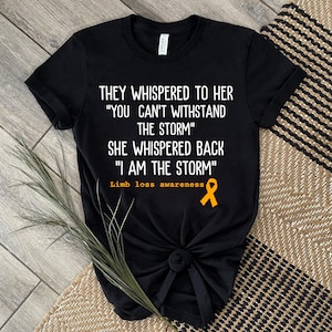 May include: Black t-shirt with white text that reads "They whispered to her "You can't withstand the storm" She whispered back "I am the storm" Limb loss awareness" and a yellow ribbon.