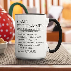 Peut inclure: Mug en céramique blanche avec une anse et un bord noirs. Le mug affiche le texte "GAME PROGRAMMER" avec une définition et le nom "Charlotte" en bas. Le mug est posé sur une surface à motifs.