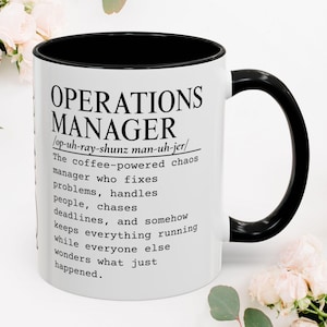 May include: A white ceramic mug with a black interior and handle, featuring black printed text that defines an Operations Manager as a coffee-powered chaos manager who fixes problems, handles people, chases deadlines, and keeps everything running.