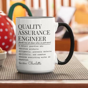 Könnte beinhalten: Weißer Keramikbecher mit schwarzem Henkel und Rand. Der Becher zeigt den Text "QUALITY ASSURANCE ENGINEER" mit einer Definition und dem Namen "Charlotte" unten. Der Becher steht auf einer gemusterten Oberfläche.