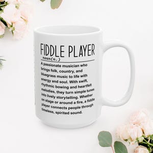 May include: White ceramic mug with the text "FIDDLE PLAYER" and a definition of a fiddle player in black font. The mug has a large handle and is surrounded by floral decorations.