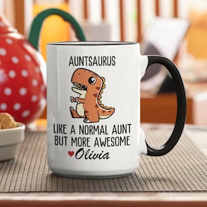 Op de afbeelding: Witte keramische mok met een zwarte rand en handvat. De mok heeft een cartoon dinosaurus met de tekst "AUNTSAURUS like a normal aunt but more awesome Olivia" met een rood hart voor de naam.