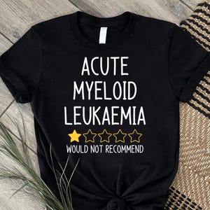 Può includere: T-shirt nera con testo bianco. Il testo recita "ACUTE MYELOID LEUKAEMIA" sopra una valutazione di una stella d'oro e quattro stelle vuote, e "WOULD NOT RECOMMEND" sotto.