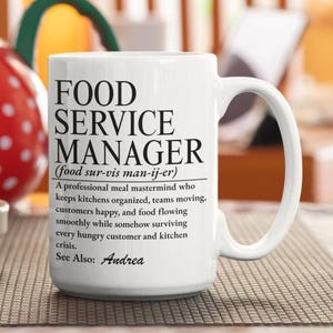 Può includere: Una tazza in ceramica bianca con testo stampato in nero che definisce un Food Service Manager come una mente professionale dei pasti che mantiene le cucine organizzate, i team in movimento e i clienti felici, con il nome Andrea.