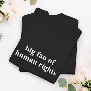 May include: Two black t-shirts are stacked. The top shirt features the white text "big fan of human rights". The shirts are on a white surface, with pink flowers in the background. The image is well-lit and the text is clear.