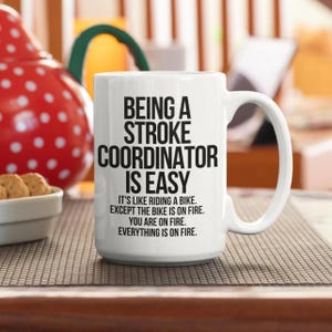 May include: White ceramic mug with the text "BEING A STROKE COORDINATOR IS EASY" in black. The mug also includes the text "IT'S LIKE RIDING A BIKE. EXCEPT THE BIKE IS ON FIRE. YOU ARE ON FIRE. EVERYTHING IS ON FIRE."
