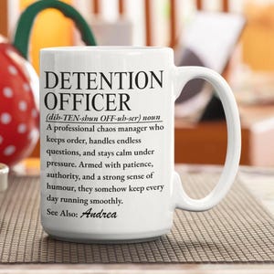 May include: A white ceramic mug featuring black printed text that defines a detention officer as a professional chaos manager who keeps order, handles endless questions, and stays calm under pressure, ending with the text See Also: Andrea.