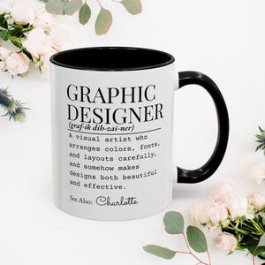Puede incluir: Una taza de cerámica blanca con interior y asa negros. El frente presenta texto negro que dice GRAPHIC DESIGNER, una pronunciación fonética, una definición del rol y el texto See Also: Charlotte en una fuente de estilo manuscrito.