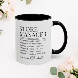 May include: White ceramic mug with a black interior and handle. The mug features the text "STORE MANAGER" with a definition, and the name "Charlotte" at the bottom. The mug is surrounded by pink flowers.