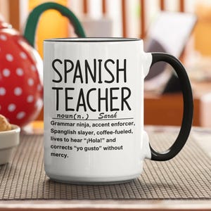 May include: White ceramic mug with a black handle and rim. The mug reads "SPANISH TEACHER" in large black letters, with a definition below. The definition includes the name "Sarah".