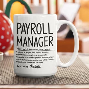 Peut inclure: Mug en céramique blanche avec l'inscription "PAYROLL MANAGER" en grosses lettres noires. En dessous, une définition et le texte "See Also: Robert". La tasse a une grande anse.