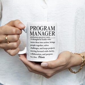 May include: A white ceramic mug featuring black printed text that defines a Program Manager as a thoughtful leader who turns ideas into action, brings people together, solves challenges, and keeps projects moving forward with clarity, collaboration, and purpose. The text also includes the name Olivia.