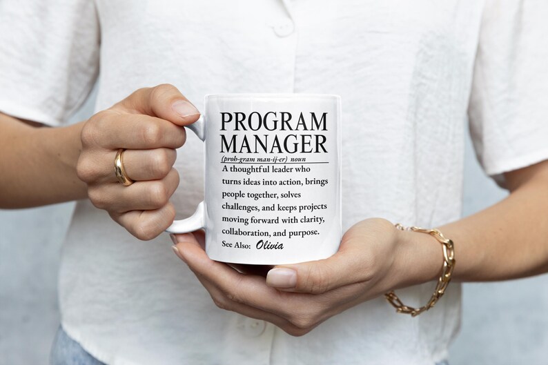 May include: A white ceramic mug featuring black printed text that defines a Program Manager as a thoughtful leader who turns ideas into action, brings people together, solves challenges, and keeps projects moving forward with clarity, collaboration, and purpose. The text also includes the name Olivia.