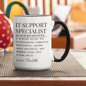 May include: White ceramic mug with a black handle and rim. The mug features the text "IT SUPPORT SPECIALIST" with a definition and the name "Charlotte" below. The mug is on a patterned surface.