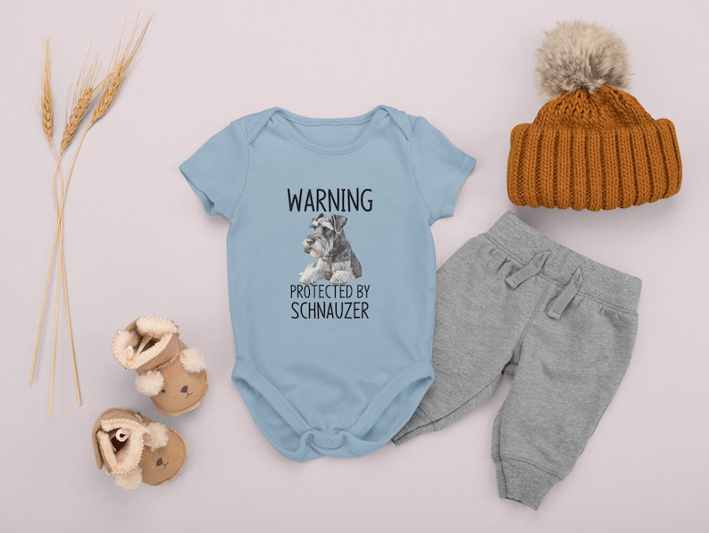 May include: A light blue baby onesie with the text "WARNING PROTECTED BY SCHNAUZER" and a picture of two miniature schnauzers. The onesie is on a white background with a brown knit hat, gray sweatpants, and brown fuzzy baby booties.