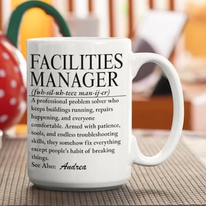 May include: A white ceramic mug featuring black printed text that defines a Facilities Manager as a professional problem solver who keeps buildings running, repairs happening, and everyone comfortable, ending with See Also: Andrea.
