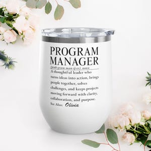 May include: A white stainless steel tumbler with a clear lid featuring black printed text that defines a programme manager as a thoughtful leader who turns ideas into action, brings people together, solves challenges, and keeps projects moving forward.