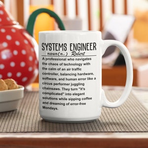 Könnte beinhalten: Eine weiße Keramik-Kaffeetasse mit einem schwarzen Design, das einen Systemingenieur als einen Profi definiert, der sich durch das Chaos der Technologie mit der Ruhe eines Fluglotsen bewegt. Die Tasse trägt den Namen "Robert" aufgedruckt.