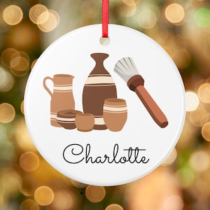 Könnte beinhalten: Ein rundes weißes Keramikornament mit rotem Bandaufhänger und einer grafischen Illustration verschiedener brauner Töpferwaren sowie einem Pinsel. Der Name Charlotte ist in schwarzer Schreibschrift unten auf dem Ornament gedruckt.