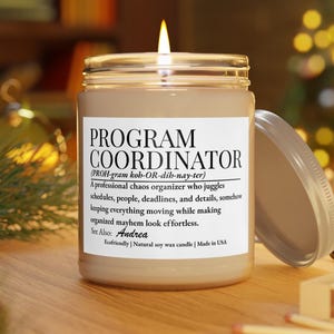 Könnte beinhalten: Eine brennende Kerze in einem klaren Glasgefäß mit einem weißen Etikett, das den Text PROGRAM COORDINATOR, eine phonetische Aussprache, eine Rollendefinition, den Namen Andrea sowie die Begriffe Ecofriendly, Natural soy wax candle enthält.