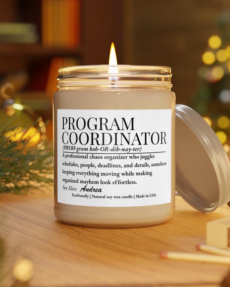 Op de afbeelding: Een brandende kaars in een doorzichtige glazen pot met een wit etiket met de tekst PROGRAM COORDINATOR, een fonetische uitspraak, een definitie van de rol, de naam Andrea en de zinsneden Ecofriendly, Natural soy wax candle en Made in USA.
