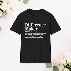 May include: Black t-shirt with the text "Difference Maker" in white, along with a definition. The definition describes someone who inspires change, uplifts others, and creates lasting impact. The shirt is surrounded by flowers.
