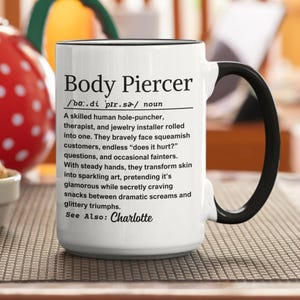 Puede incluir: Taza de cerámica blanca con asa y borde negros. La taza presenta el texto "Body Piercer" con una definición. La descripción incluye el texto "See Also: Charlotte".