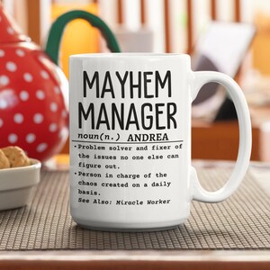 Puede incluir: Taza de cerámica blanca con un diseño negro que dice "MAYHEM MANAGER noun(n.) ANDREA • Problem solver and fixer of the issues no one else can figure out. • Person in charge of the chaos created on a daily basis. See Also: Miracle Worker"