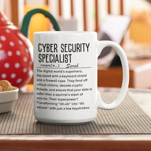 Op de afbeelding: Een witte keramische mok met een zwart ontwerp met de tekst "Cyber Security Specialist" en definieert de term als "De superheld van de digitale wereld, uitgerust met een toetsenbord schild en een firewall cape. Ze weren virtuele schurken af, decoderen cryptische bedreigingen en zorgen ervoor dat uw gegevens veiliger zijn dan de voorraad eikels van een eekhoorn. Hun superkracht? "Uh-oh" transformeren in "Alles veilig" met slechts een paar toetsaanslagen." De naam "Sarah" is opgenomen in de definitie.