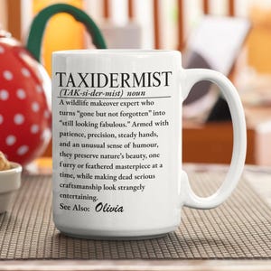 Pode incluir: Uma caneca de cerâmica branca com texto impresso em preto que define a palavra TAXIDERMIST como um especialista em transformação da vida selvagem. O texto inclui pronúncia fonética, uma descrição humorística e o nome Olivia sob See Also.