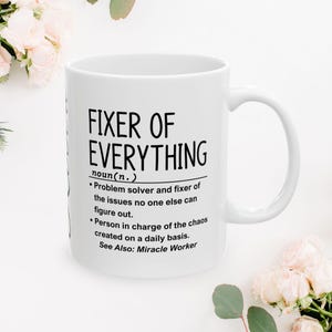 Könnte beinhalten: Weiße Keramik-Tasse mit dem schwarzen Text "FIXER OF EVERYTHING". Die Tasse enthält eine Definition des Begriffs, die einen Problemlöser und jemanden beschreibt, der für das tägliche Chaos verantwortlich ist. Die Tasse ist von weißen Blumen umgeben.