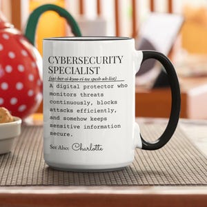 May include: A white ceramic mug with a black handle and rim. The mug features the text "CYBERSECURITY SPECIALIST" and a definition, along with the name "Charlotte". The mug is on a patterned surface.