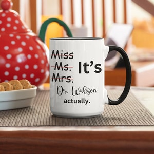May include: White coffee mug with a black rim and handle. The mug has black text that reads "Miss Ms. Mrs. Dr. Wilson It's actually."