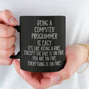 May include: Black coffee mug with white text that reads "Being a Computer Programmer is Easy. It's like riding a bike. Except the bike is on fire. You are on fire. Everything is on fire."
