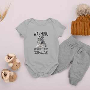 May include: A gray baby onesie with a white graphic that says "WARNING PROTECTED BY SCHNAUZER" and a picture of two miniature schnauzers. The onesie is paired with gray sweatpants and a brown knit beanie with a white pom-pom.