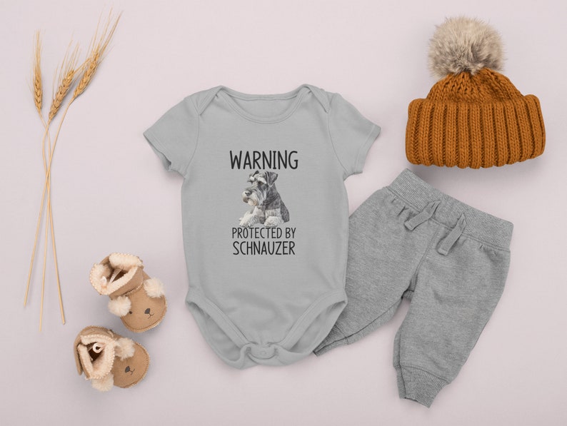 May include: A gray baby onesie with a white graphic that says "WARNING PROTECTED BY SCHNAUZER" and a picture of two miniature schnauzers. The onesie is paired with gray sweatpants and a brown knit beanie with a white pom-pom.