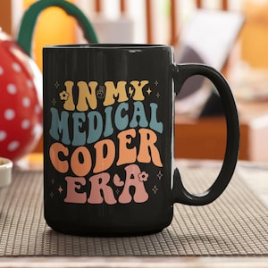 May include: A black ceramic mug featuring the text "IN MY MEDICAL CODER ERA" in a multi-colored, wavy font. The design includes small flower and star icons interspersed among the words, with the lettering in shades of yellow, blue, orange, and pink.